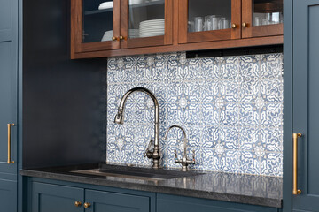 A kitchen sink with a beautiful pattern tiled backsplash with a chrome faucet, black granite...