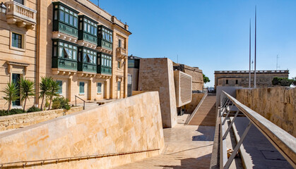 A historic Maltese house and a modern Parliament building with city gate stairs and walls,...