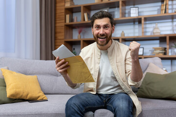 Portrait of happy man at home sitting on sofa rejoicing and celebrating victory and success looking at camera and holding hand up gesture of triumph, man received envelope mail letter with good news. - obrazy, fototapety, plakaty