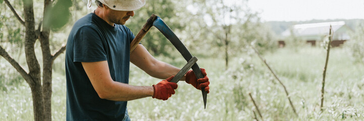 mowing grass traditional old-fashioned way with hand scythe on household village farm. young mature farmer man sharpening scythe with grass or whetstone for mowing the grown weed of a farmland. banner