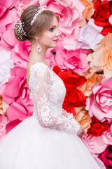A girl in a white wedding dress with an open back, make-up, a hairstyle in the form of a bun poses on a bright red background of paper flowers.