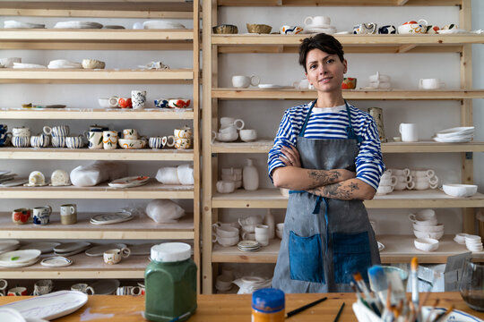 Creative ceramics studio owner at workplace standing arms crossed and looking at camera. Confident entrepreneur crafts woman in pottery studio preparing for master class or work process