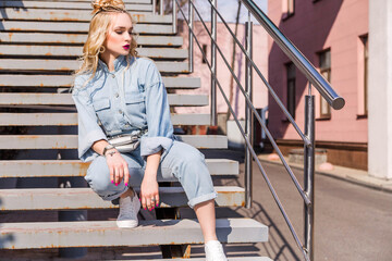 A blonde girl, with bright make-up and a hairstyle in denim overalls, sits on the stairs on the street.