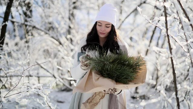 Happy young woman stands in winter forest with bouquet from pine branches in her hands.