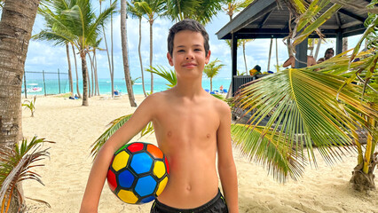 Portrait of a cute European teen boys wearing swimming shorts. The boy standing on the beach holding a ball. © Danko