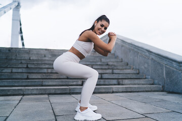 Young woman makes squats to pump the muscles of buttocks and make fit form, simple and effective exercises during morning workout in city. Healthy lifestyle, female keeping the body in sporty shape