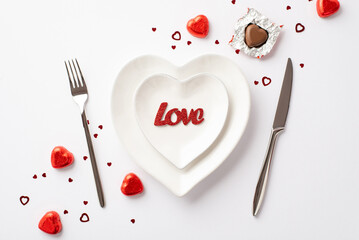 Valentine's Day concept. Top view photo of heart shaped plates with inscription love knife fork chocolate candies and confetti on isolated white background