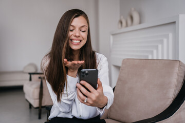 Naklejka premium Cheerful Hispanic young woman holds phone sends air kiss during video call sitting on cozy chair at home dressed in white shirt. Pretty brunette businesswoman grateful to her husband. Happy woman.