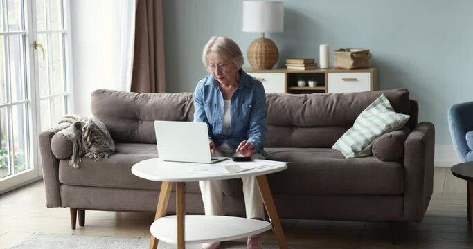 Smiling older woman wear glasses sit at table with laptop in living room, calculates household utility, managing family budget feels satisfied having enough of pension to pay bills. Finances control