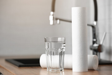 A glass of clean water and foamed polypropylene filter cartridges on wooden table in a kitchen interior. Installation of reverse osmosis water purification system. Concept Household filtration system.