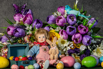 toddler doll sitting in front of spring flowers with stuffed rabbit jelly beans and colored Easter eggs