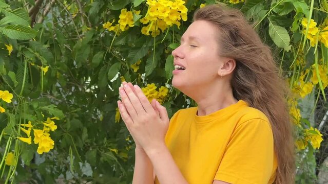Portrait of beautiful young allergic woman is suffering from pollen allergy or cold on natural flower flowering tree background at spring or sunny summer day sneezes, blowing her runny nose rubs eyes