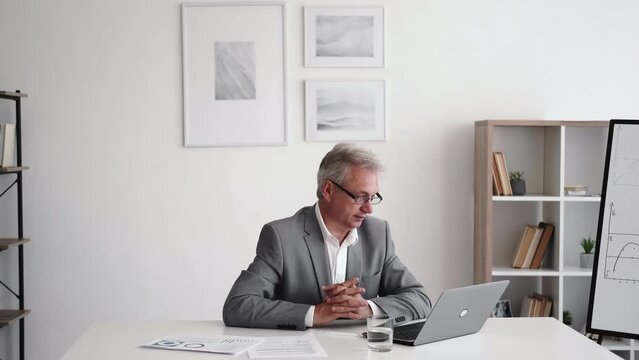 Business webinar. Video interview. Corporate telecommuting. Confident smart senior male coach discussing success strategy using laptop at light office interior with free space.
