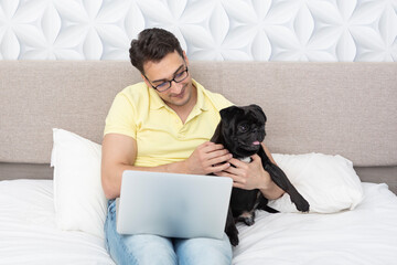 Smiling happy man sitting on the bed and hugging his lovely pet black pug breed and working on laptop. Friendship concept