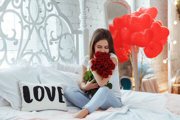 Happy girl with red roses on bed and air balloons in shape of heart on the background. Celebrating Saint Valentines day.