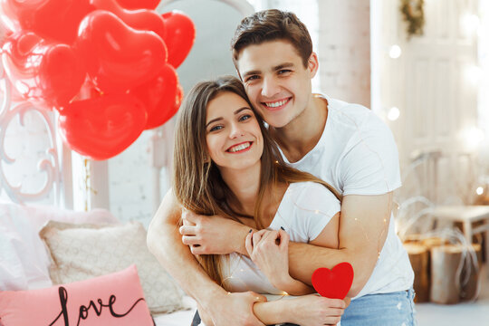 Beautiful young couple at home. Hugging, kissing and enjoying spending time together while celebrating Saint Valentine's Day with air balloons in shape of heart on the background.