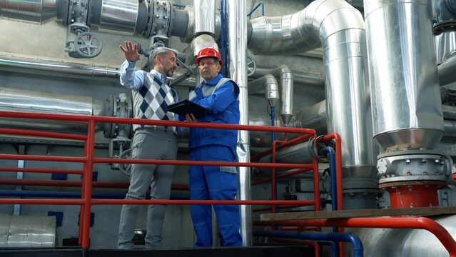 Production Manager Controls the Work of an Employee in District Heating Plant. Factory Interior with Large Piping. Global Energy Crisis. Supervisor Using Digital Tablet and Talking to Worker.