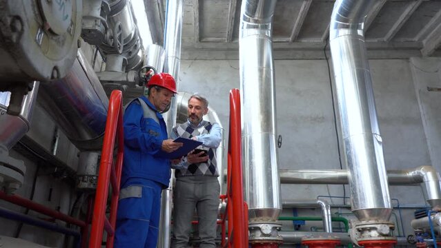 Production Manager Using Digital Tablet and Talking to Worker in District Heating Plant. Digital Technology and Teamwork Concept. Industry 4.0. Factory Interior with Large Piping. Global Energy Crisis