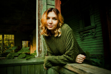 Portrait of a beautiful blonde woman in a green vintage sweater in an old wooden abandoned house overgrown with plants in the summer.