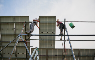 View from below of two men solar technicians standing on metal rail and mounting photovoltaic solar panel. Male workers installing solar modules under white blue sky. Concept of sustainable energy.