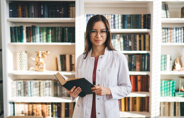 Positive medical worker standing with book in library - obrazy, fototapety, plakaty