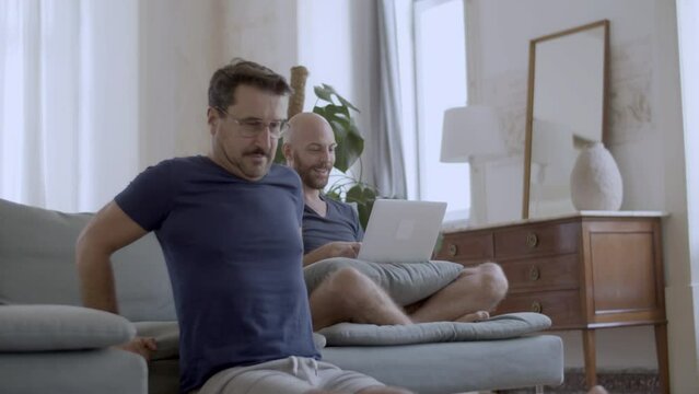 Handsome gay in eyeglasses doing push-ups off couch at home. Happy homosexual man sitting on sofa with laptop while his partner doing physical exercises and smiling. Sport, love, LGBT concept