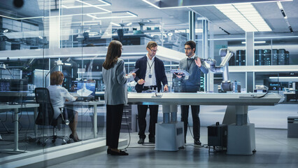 Diverse Team of Industrial Robotics Engineers Gathered Around a Table With Robotic Arm. Scientists Use Tablet Computer to Manipulate and Program the Robot to Pick Up and Move a Metal Component.