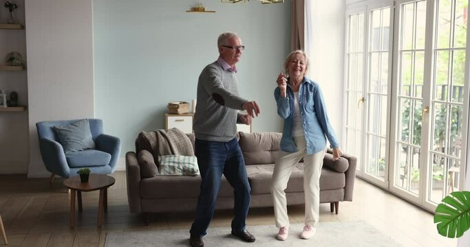 Carefree couple of retirees, older wife and husband dancing in living room, enjoy music and wellbeing spend active leisure together at modern cozy home. Hobby, lifestyle, untroubled life of seniors