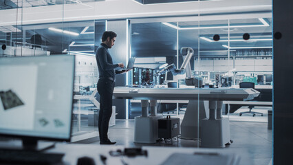 Industrial Robotics Engineer Standing Next to Robotic Arm in a High Tech Factory. Male Scientist Uses Laptop Computer to Manipulate and Program the Robot to Pick Up and Move a Microchip.