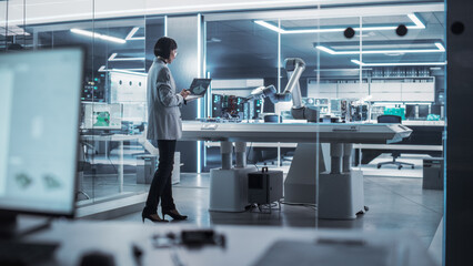 Industrial Robotics Engineer Standing Next to a Robotic Arm in a High Tech Factory. Female Scientist Uses Laptop Computer to Manipulate and Program the Robot to Pick Up and Move a Microchip.