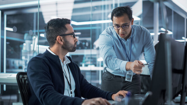 Two Industrial Engineers Discussing Work Projects, Talk and Use Desktop Computer in Robotics Startup Facility. Specialists and Professionals Researching and Developing High Tech Robotics Equipment.