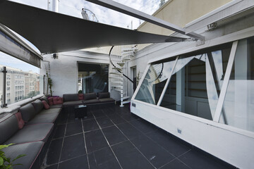 Terrace of an attic house with built-in seats with cushions in a large part of the perimeter, a spiral staircase and glass partitions