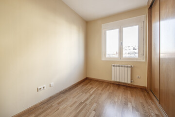 Fototapeta na wymiar Room with a built-in wardrobe with three sliding oak doors with skirting boards and matching floors and smooth cream-colored walls and a white aluminum window with two hinged leaves