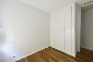 Room with a white wooden four-door built-in wardrobe, a dark wooden floor and plain white walls