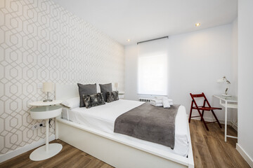 Bedroom with a white wooden headboard, gray cushions, a white wooden bed and in a corner a desk with a red wooden folding chair - Powered by Adobe