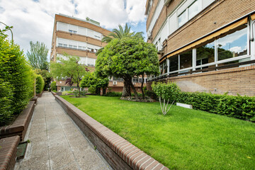 Facade of a four-story building with white aluminum windows and walkways with lawn and tree-lined gardens