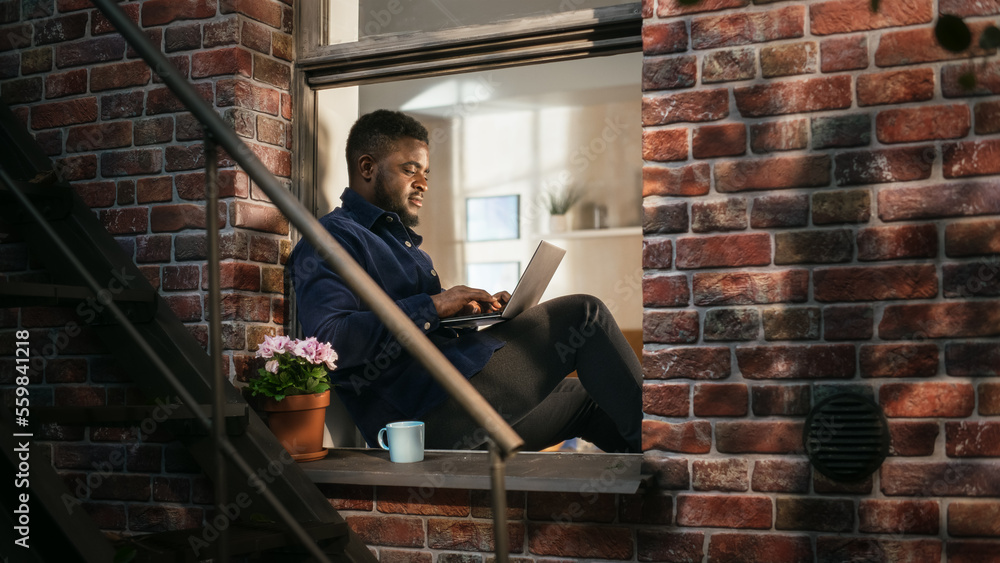Poster Black Young Male Smiling and Using Laptop While Sitting on a Windowsill. African American Businessman Enjoying the Weather While Working from Home. He Is Thinking and Answering his Business Emails.