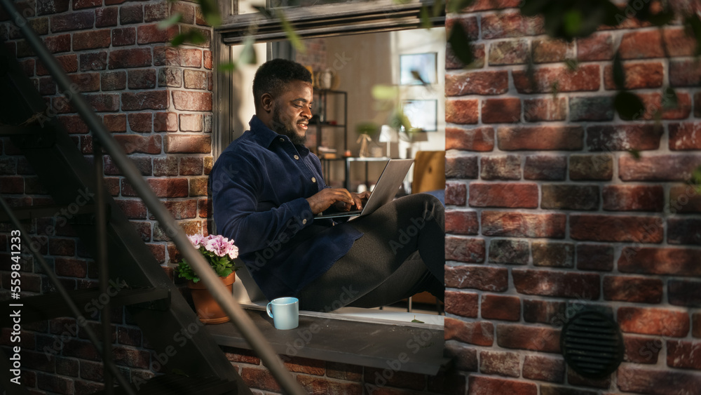 Poster smiling black man working on laptop computer while sitting on a windowsill in a stylish cozy brookly
