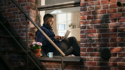 Black Young Male Smiling and Using Laptop While Sitting on a Windowsill. African American...