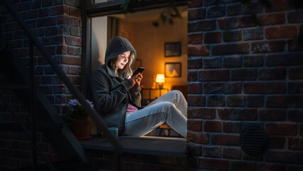 Focused Woman Typing Messages on her Smartphone While Sitting By a Window in the Dark. Internet Addicted Female Using Mobile Phone App for Online Shopping, Social Media Posting, Internet Browsing