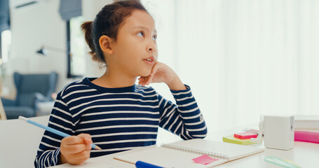 Fototapeta na wymiar Asian toddler girl with sweater sit in front of desk with notepad use pencil focus on write notebook do homework from online learning course on the weekend at home. Distance online learning concept.