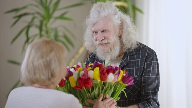 Loving senior husband passing bouquet of flowers to adorable wife in slow motion talking smiling. Portrait of confident handsome Caucasian man congratulating woman on Valentine's Day