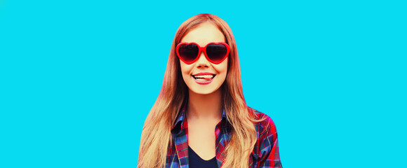Portrait of happy smiling young woman teasing wearing red heart shaped sunglasses on blue background
