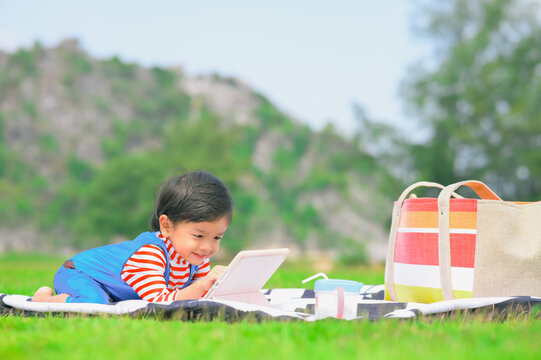 Asian Kid Boy Enjoying Laying Down On Picnic Blanket In Park,using Tablet ,sunny Green Park Outdoors. Active Weekend Leisure Recreation