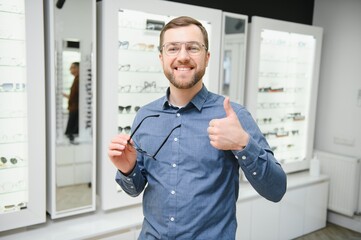 Satisfied Customer. View of happy young male client wearing new glasses, standing near rack and showcase with eyewear. Smiling man trying on spectacles