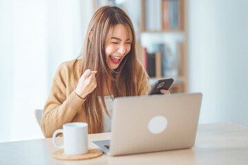 Fototapeta premium Excited young asian woman winner looks at laptop celebrates online work at home on desk with successful. Reads good news in email, Achieve target project.