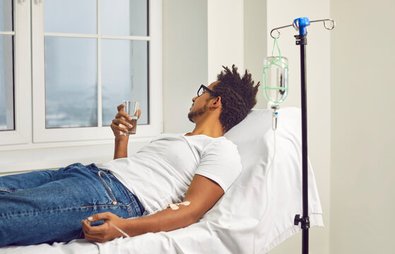 Portrait of a sick young african american man in the glasses lying in a white medical ward under a dropper and drinking a glass of water looking out the window. Healthcare and medicine concept.