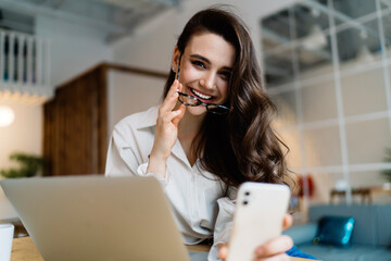 Obraz premium Happy female blogger talking selfie on smartphone sitting at table with laptop