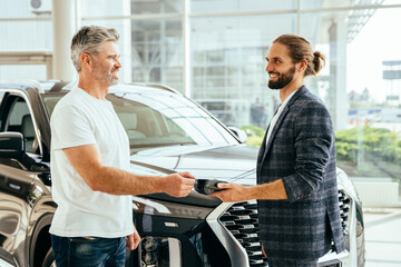 Handsome grey hair mature man in white t-shirt buying a car, paying using a terminal with a bank card in a modern car dealership. Successful man buying brand new luxury car in showroom.