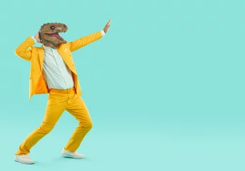 Foto op Plexiglas Carnaval Strange guy in reptile costume dancing and having fun. Crazy young man wearing yellow suit and funny dinosaur mask dancing and moving hands isolated on blank turquoise copy space background